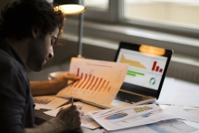 Man looking at computer screen and many sheets of graphs and data