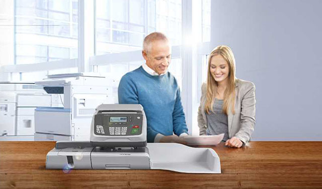 A blond woman in a grey blazer and a man wearing a blue sweater standing next to a postage meter in a bright office space with large windows in the background
